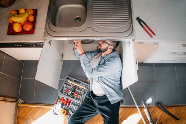 Man fixing plumbing under a kitchen sink with tools spread out.